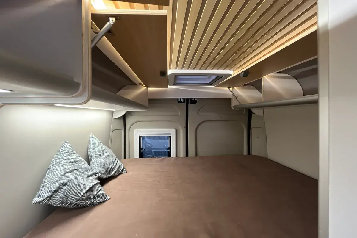 Zona de dormir en una camper de alquiler con cama de matrimonio, almohadas y techo de madera